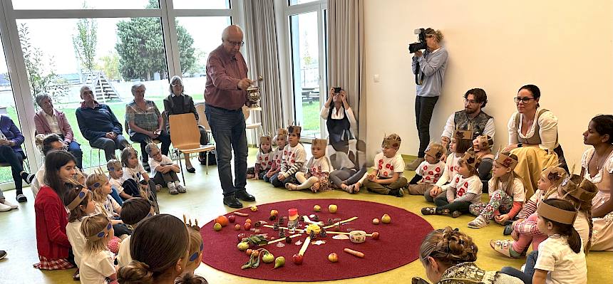 Kinder sitzen im Sitzkreis, Pfaffer segnet